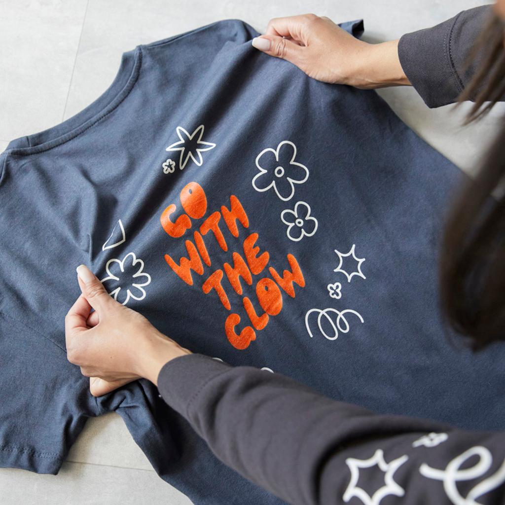 How to start a print on demand business on Etsy Hands hold a dark blue t-shirt featuring the phrase "GO WITH THE GLOW" in bold orange letters, surrounded by white floral designs.