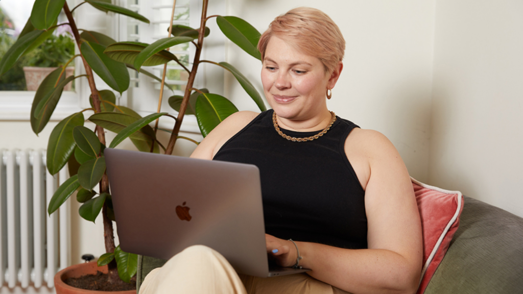 Printify Pop Up Store – What is its biggest perk A woman with short hair smiles while working on a laptop. She sits on a couch beside a potted plant.